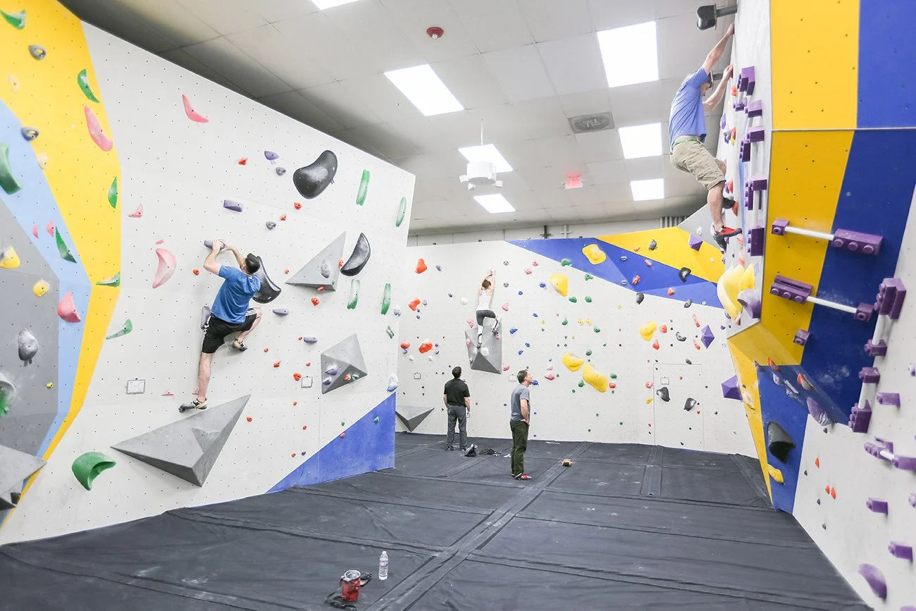 Indoor rock climbing gym with various colored climbing holds on white, yellow, and blue walls. Several climbers are actively climbing, and a couple of people are standing and observing at the bottom.
