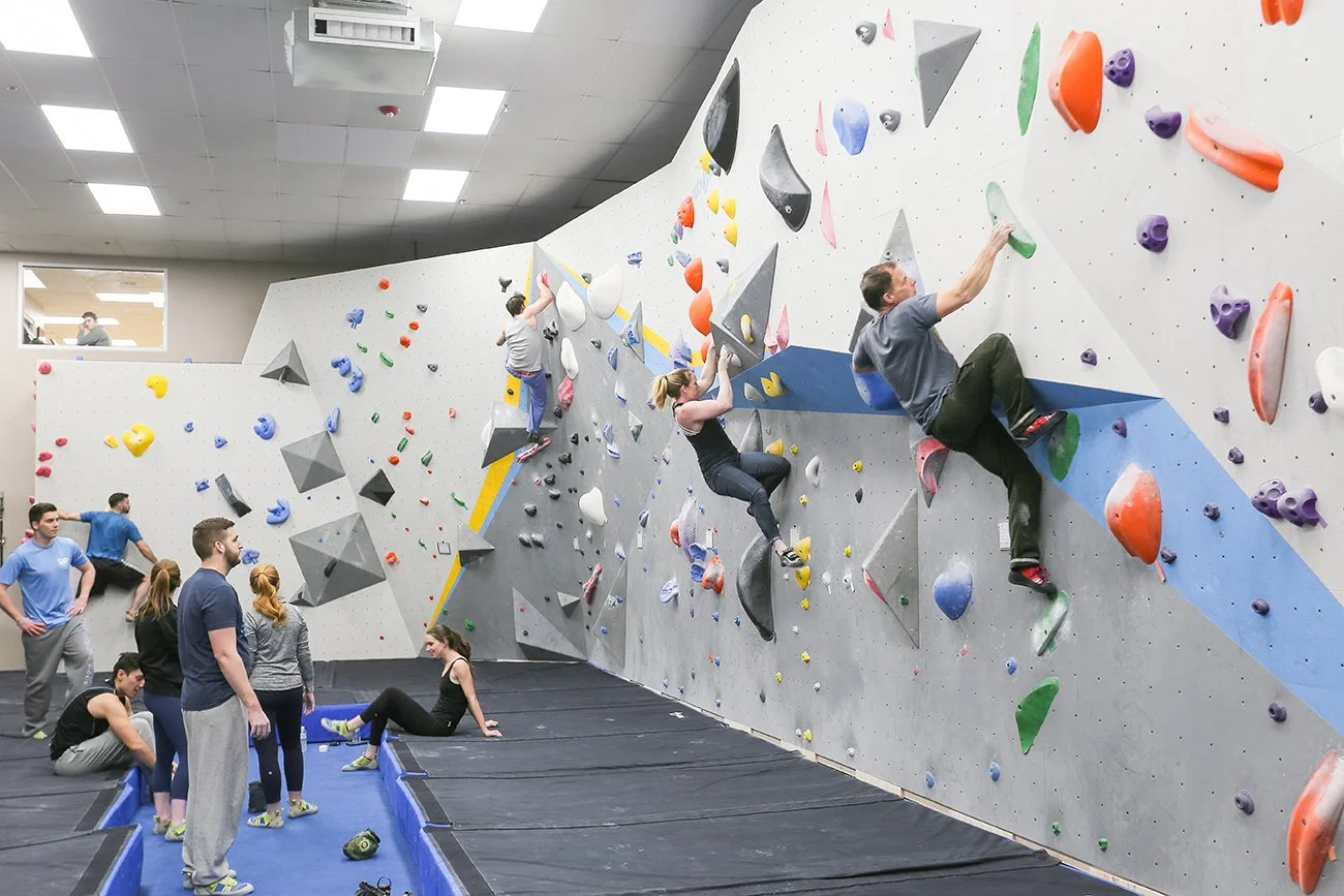 People climbing on an indoor bouldering wall; others watching or resting nearby.