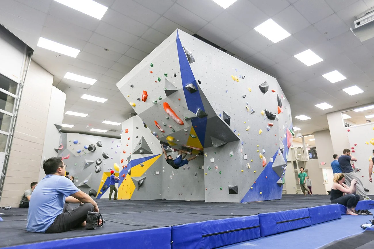 Indoor climbing gym with bouldering walls, climbers, and spectators sitting on padded flooring.