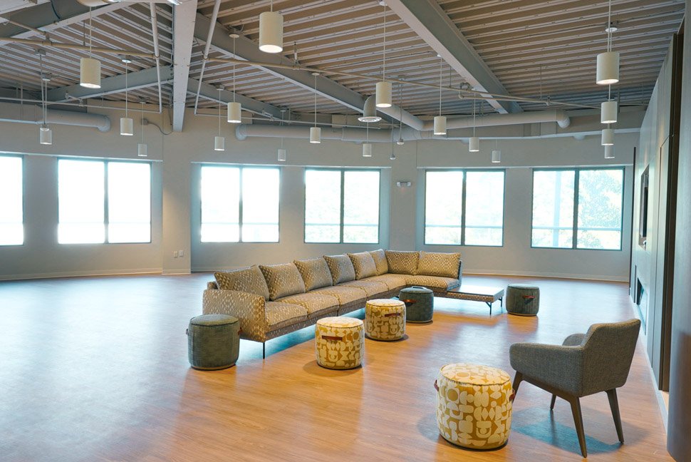 A spacious, modern lounge area with a long beige couch, several patterned ottomans, and a single gray armchair, illuminated by large windows and ceiling lights, with a wood floor and exposed ceiling beams.