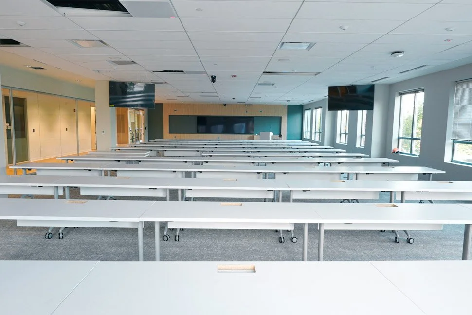 Empty classroom or conference room with long white tables, multiple black monitors, and large windows letting in natural light.