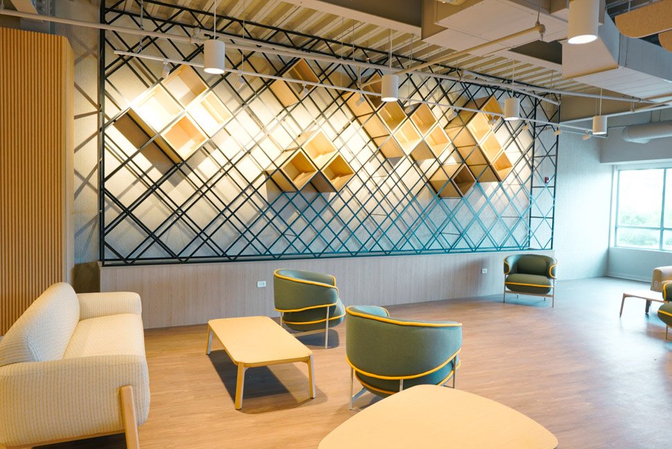 Modern office lounge with green and beige chairs, a wooden coffee table, and a geometric wall decor with wooden shelves and black metal frame.