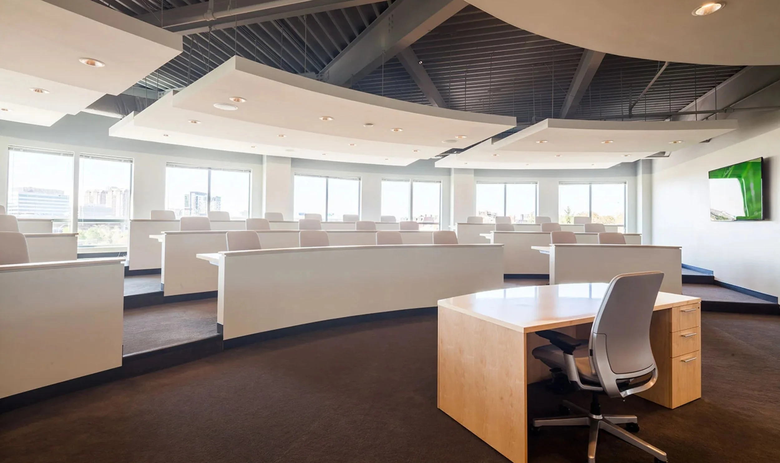 Empty office classroom with desks and chairs facing large windows, a television on the wall, and office desk at the front.