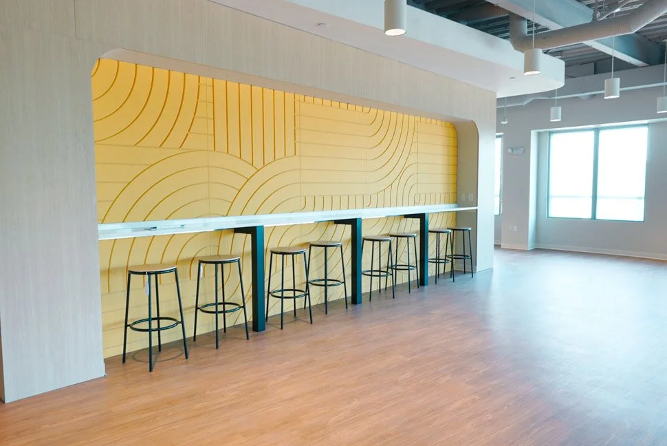 Empty modern room with a bar-height counter and six black stools, yellow decorative wall with curved line patterns, and large windows letting in natural light.
