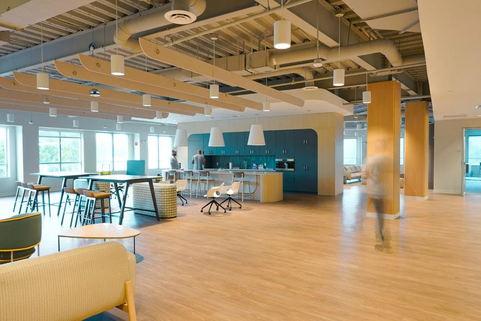 Modern, open-concept office lounge with high tables, bar stools, a kitchenette area, large windows, and wooden accents.