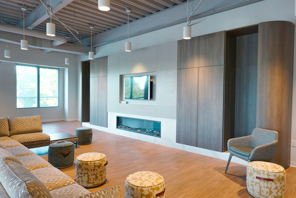 Modern living room with a large beige sectional sofa, small round ottomans, a gray armchair, a wall-mounted TV, a fireplace, a window, and contemporary ceiling lights.
