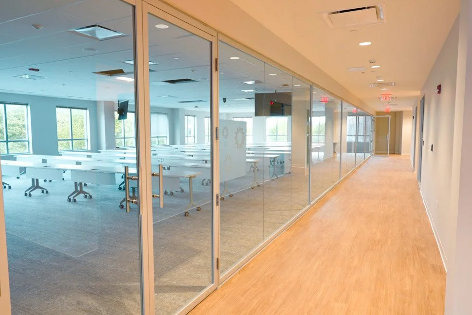 Empty classroom with white desks and chairs, glass walls, and windows showing trees outside.
