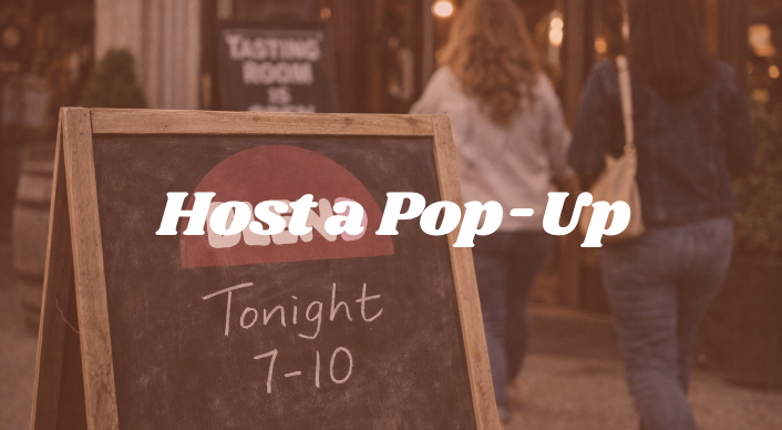 Outdoor chalkboard sign promoting a pop-up event tonight at a Woodinville wine tasting room from 7 to 10, with people walking nearby.