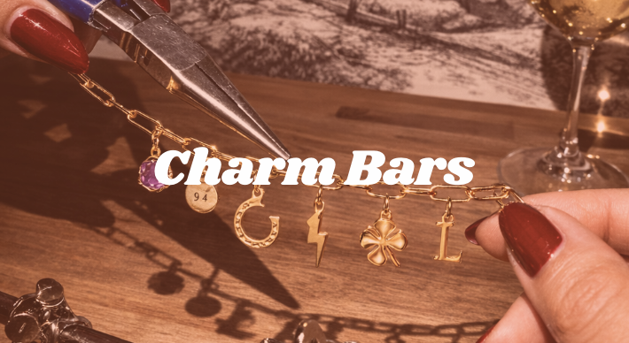 Hand holding a charm bracelet with various gold charms, hand stamped disc, crescent moon, lightning bolt, four-leaf clover, and a letter L, with the words "Charm Bars" overlayed. The background features a wooden surface, a pen, and a glass of wine.