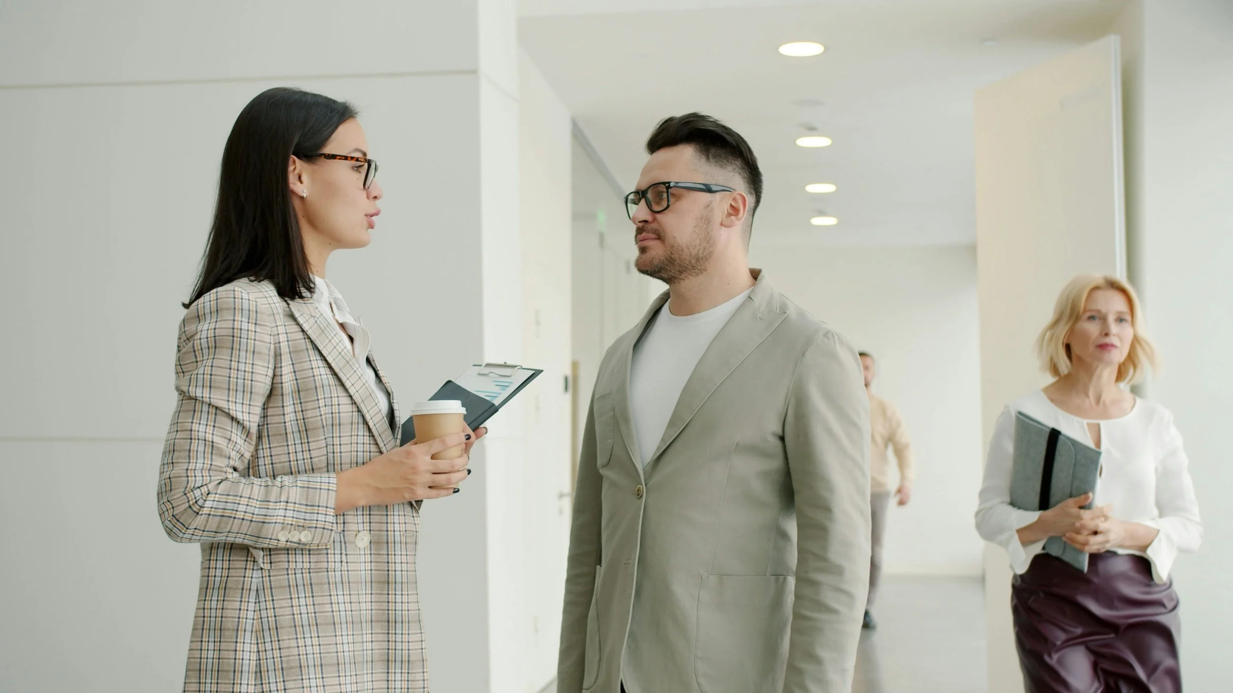 A woman with long dark hair holding a coffee cup and tablet, wearing glasses and a plaid blazer, talking to a man with short dark hair and glasses, wearing a light-colored blazer and white shirt, in a bright hallway with other people visible in the background.