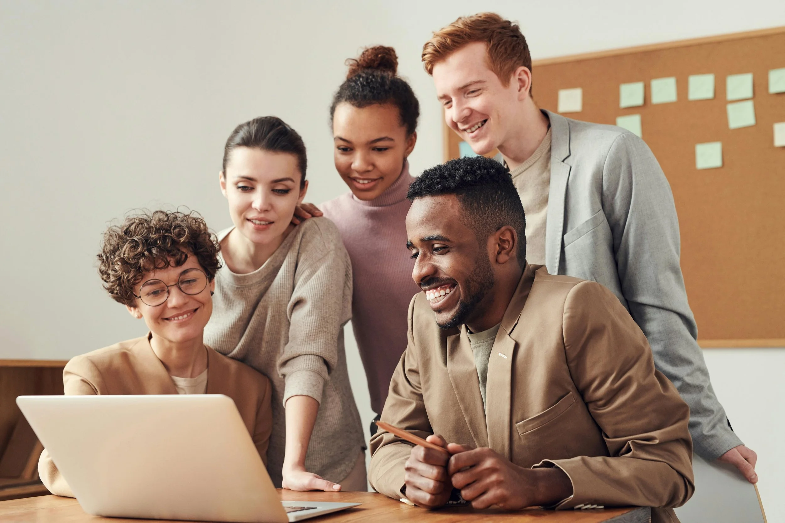Six people gathered around a laptop, smiling and looking at the screen.
