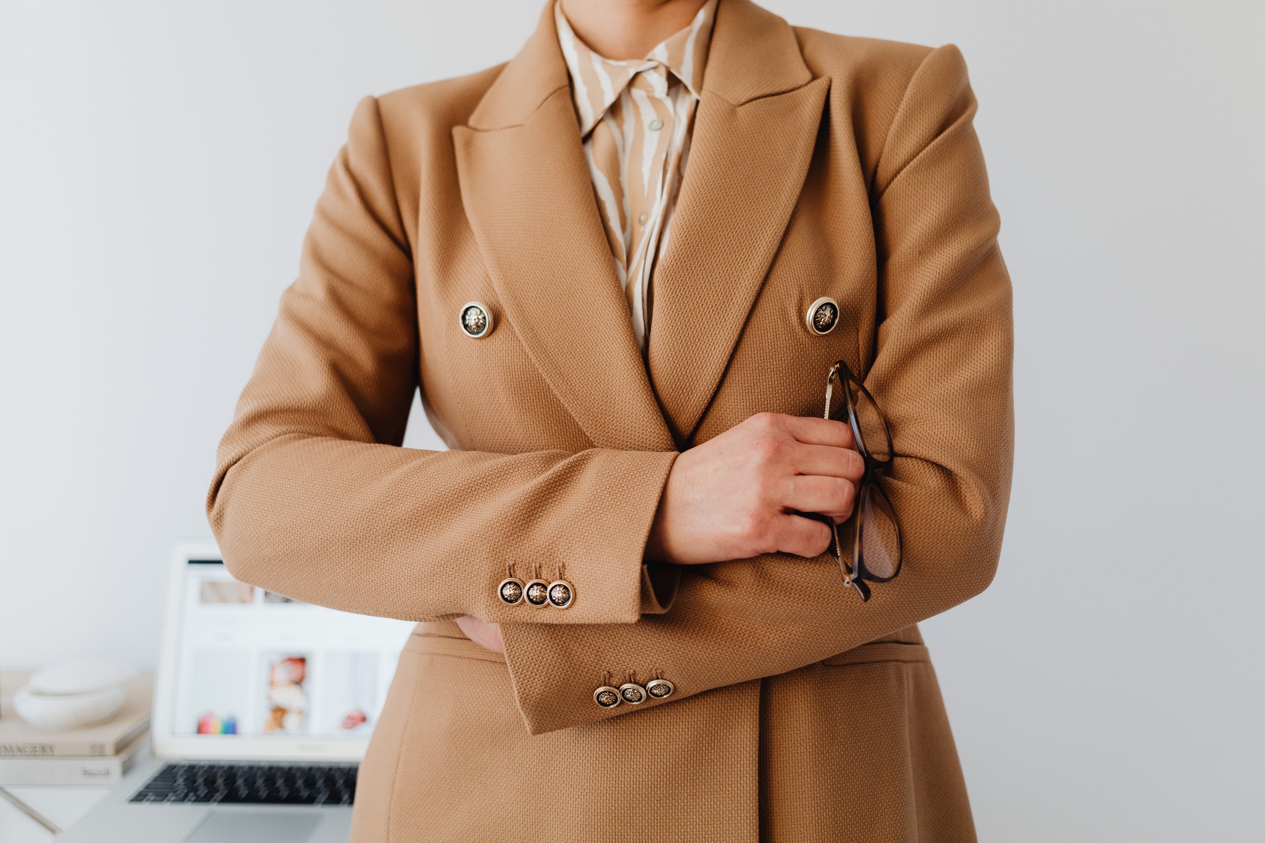 Person wearing a tan blazer with gold buttons, holding eyeglasses, with a laptop and books in the background.