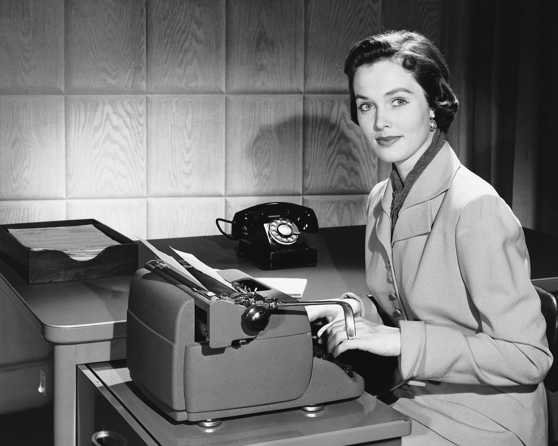 A woman in professional attire sitting at a desk, typing on a vintage typewriter with a rotary phone and files nearby, in a black-and-white photo.