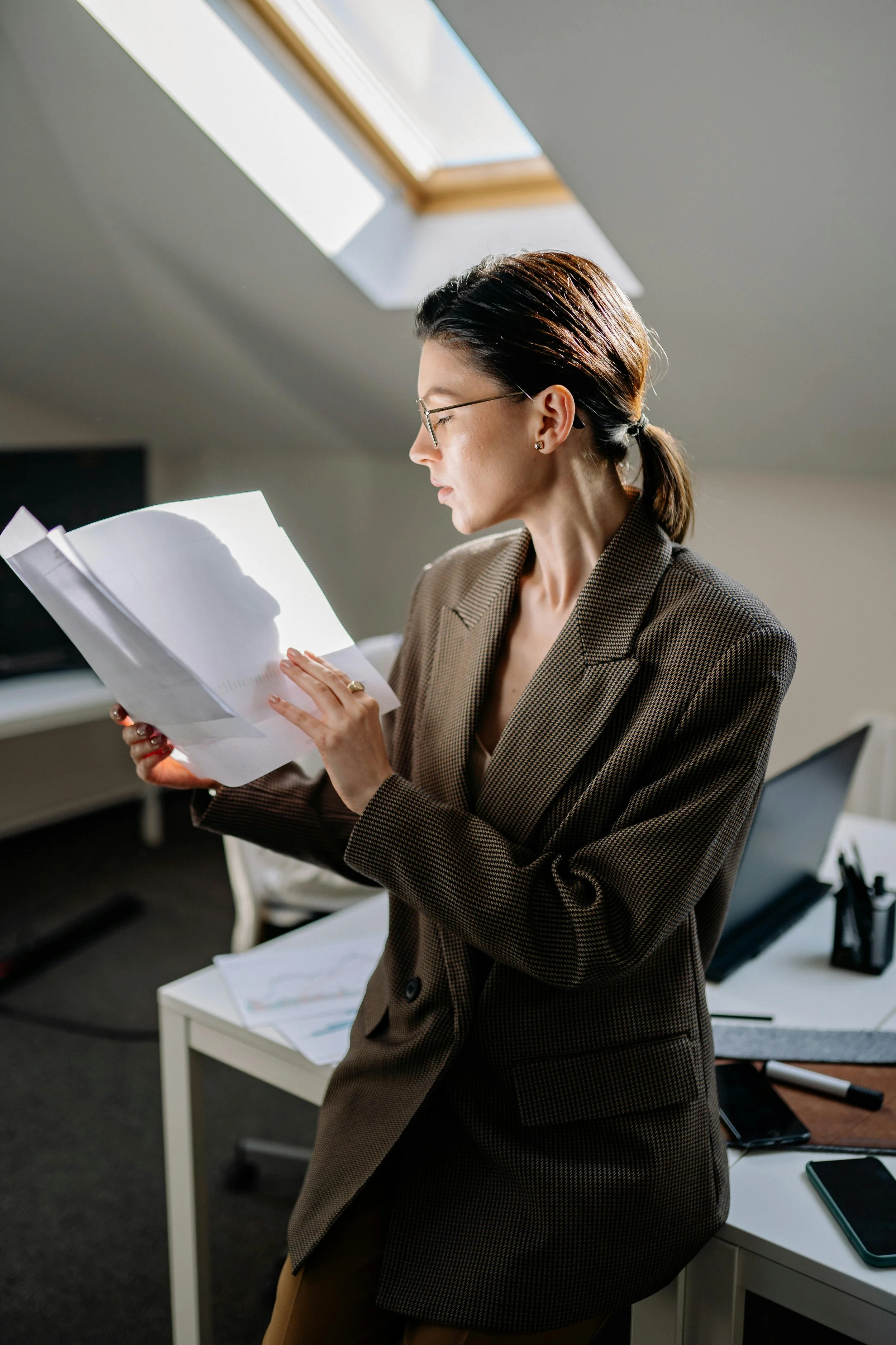 A woman wearing glasses and a brown blazer, standing in an office with a slanted ceiling and a skylight, reading a document.