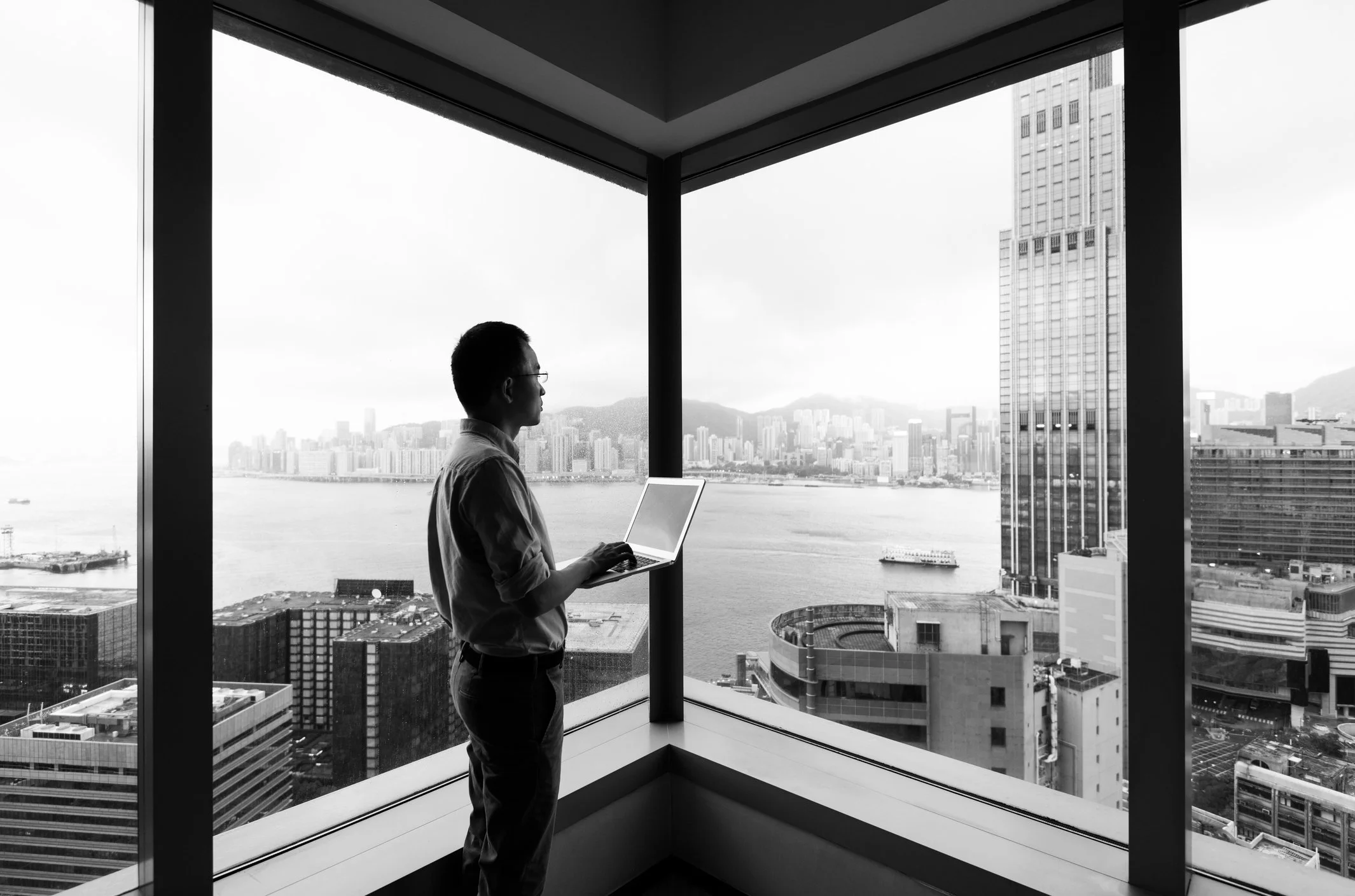 A man in business attire working on a laptop in a high-rise office with large windows, overlooking a cityscape with water, boats, and tall buildings.