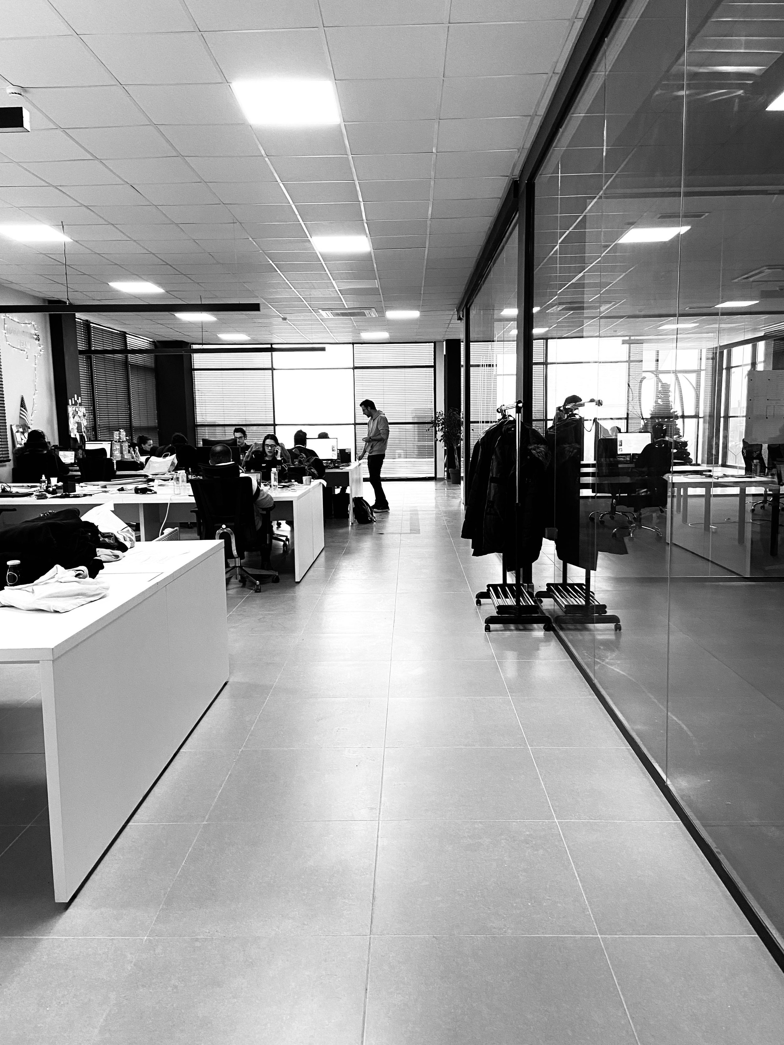 An office space with people working at desks, a man standing, and a clothing rack with jackets near a glass wall.