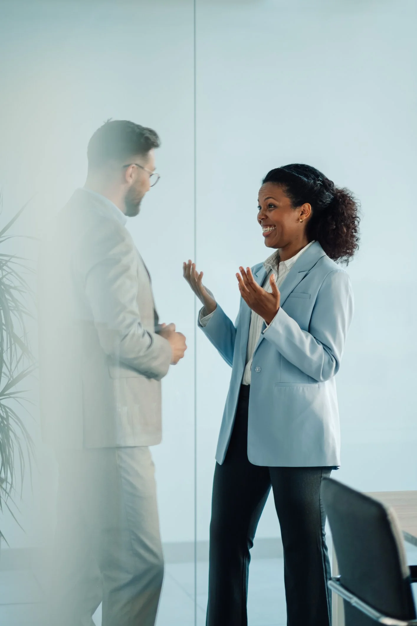 Two professionals, a man and a woman, are engaging in a conversation in an office setting. The woman is smiling and gesturing with her hands.