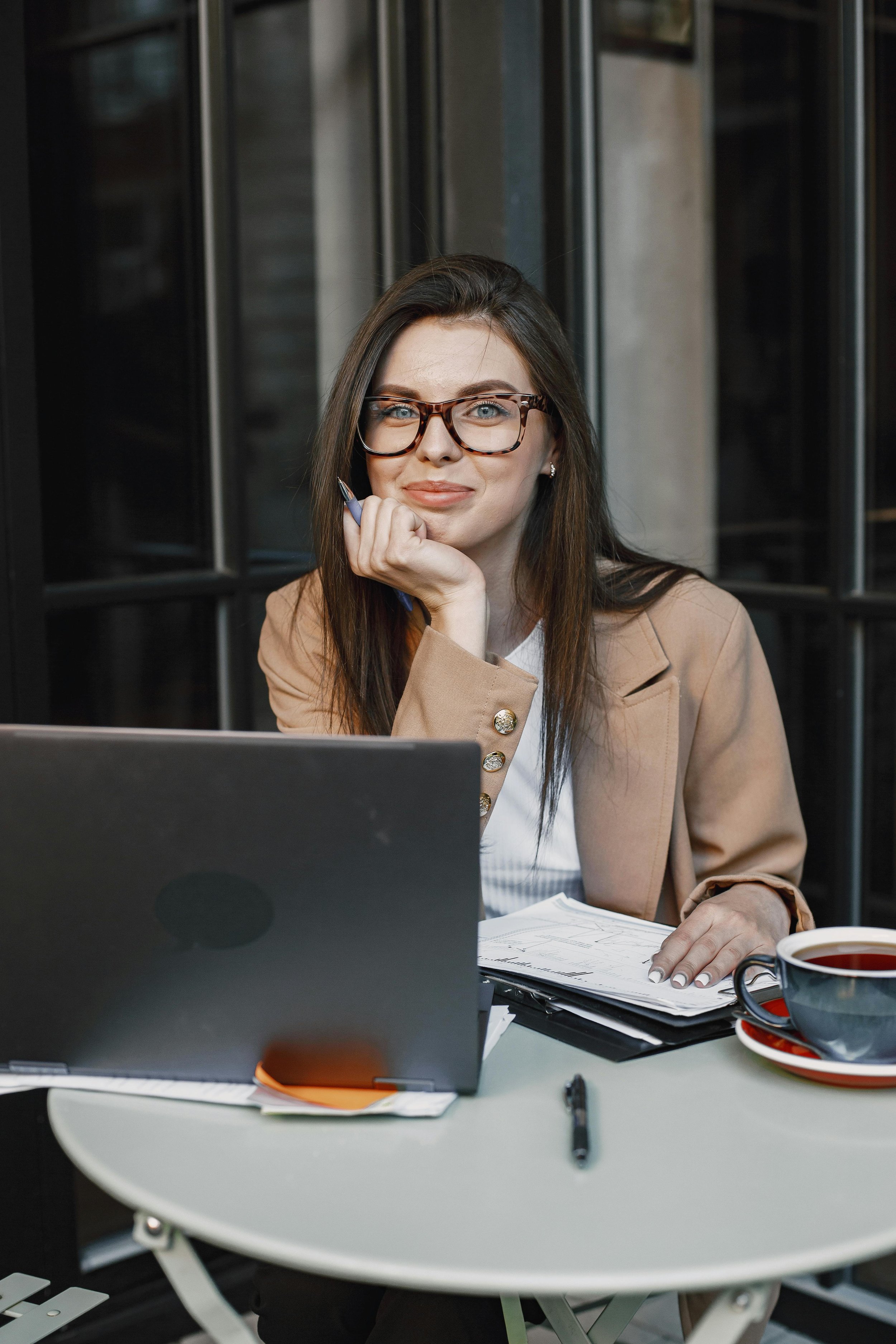A young woman with long brown hair and glasses sitting at a table with a laptop, cup of tea, and documents, resting her chin on her hand and looking at the camera.