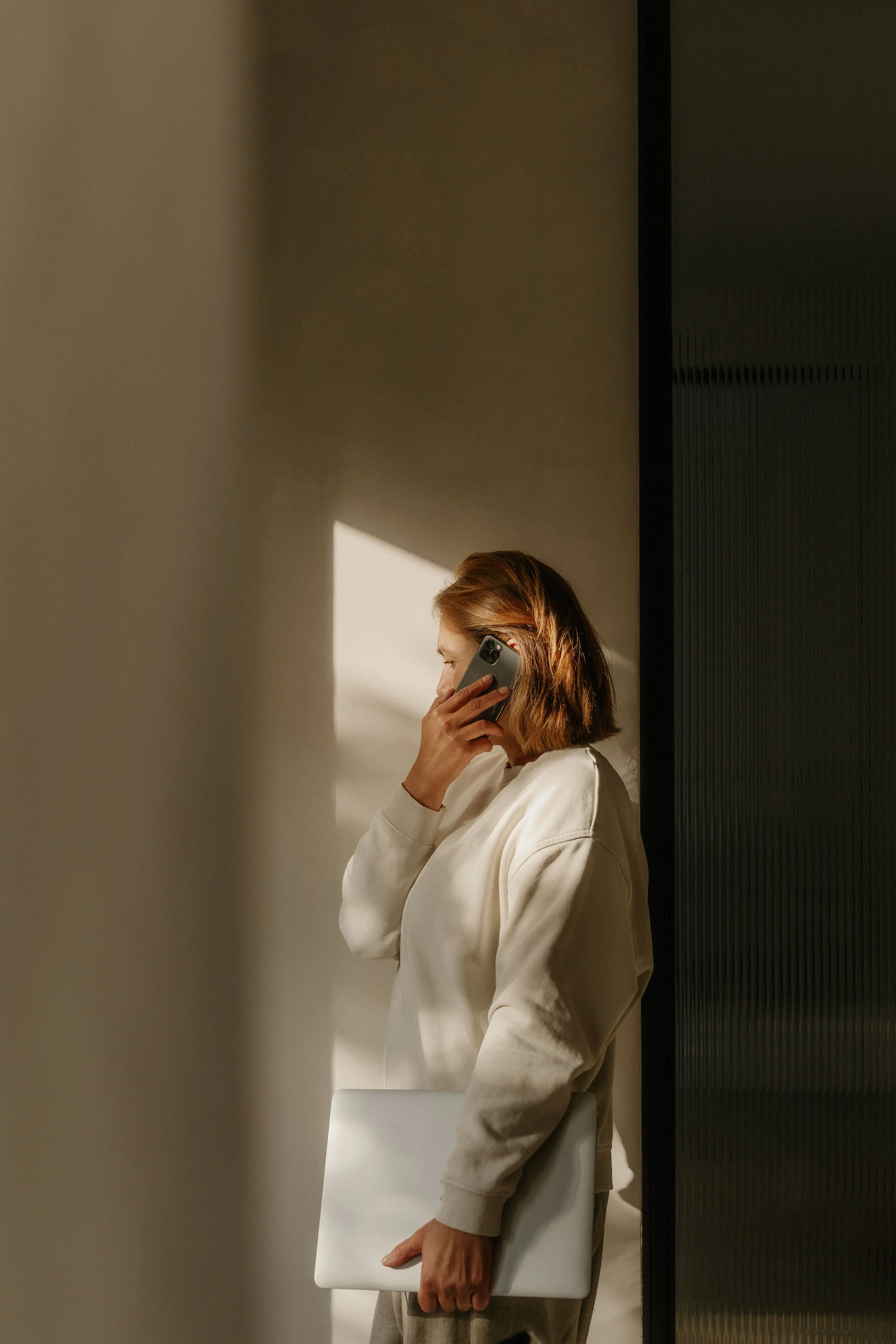 A woman with shoulder-length red hair, wearing a beige sweatshirt, is holding a smartphone to her ear and a closed laptop in her other hand. She is standing near a wall with sunlight casting a shadow on the wall behind her.