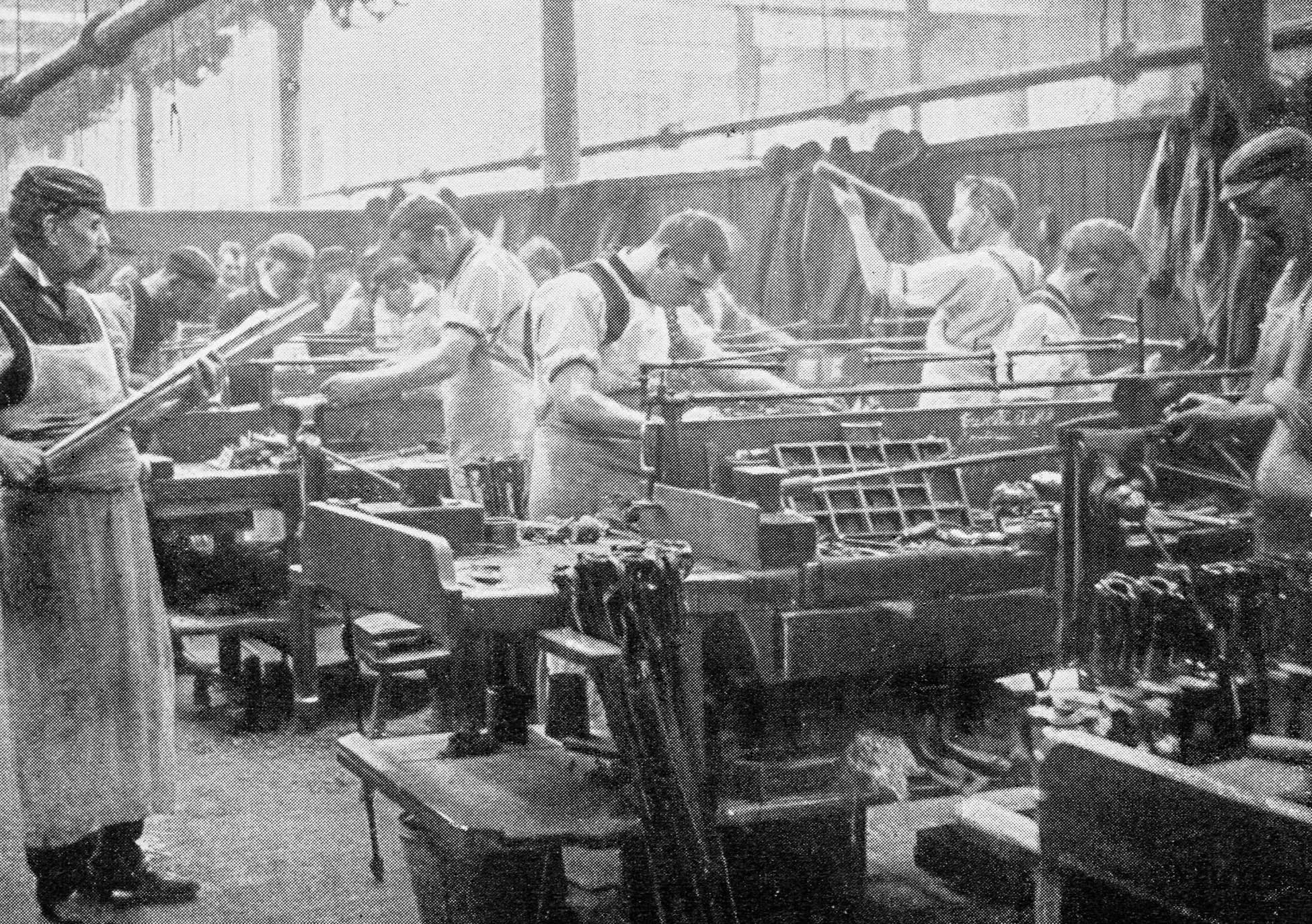A group of women working on assembly line assembling toy trains, wearing aprons and hair coverings in a factory setting.