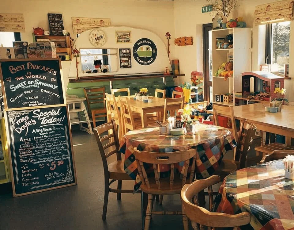 Cozy cafe interior with wooden tables and chairs, decorated with colorful checkered tablecloths, small plants, and condiments. A chalkboard menu displays breakfast options, and a white shelf holds stuffed animals and books near sunlight-filled windows.