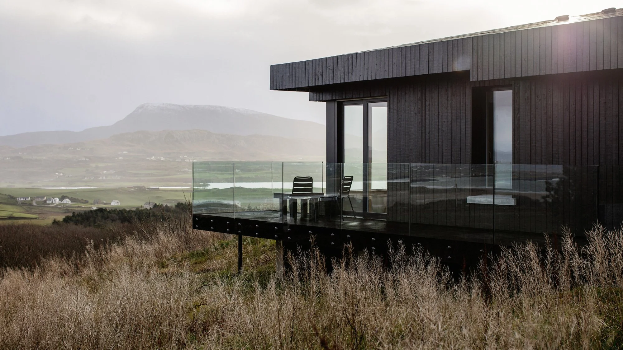 A modern black house with a glass balcony overlooking a grassy field and mountains in the distance.