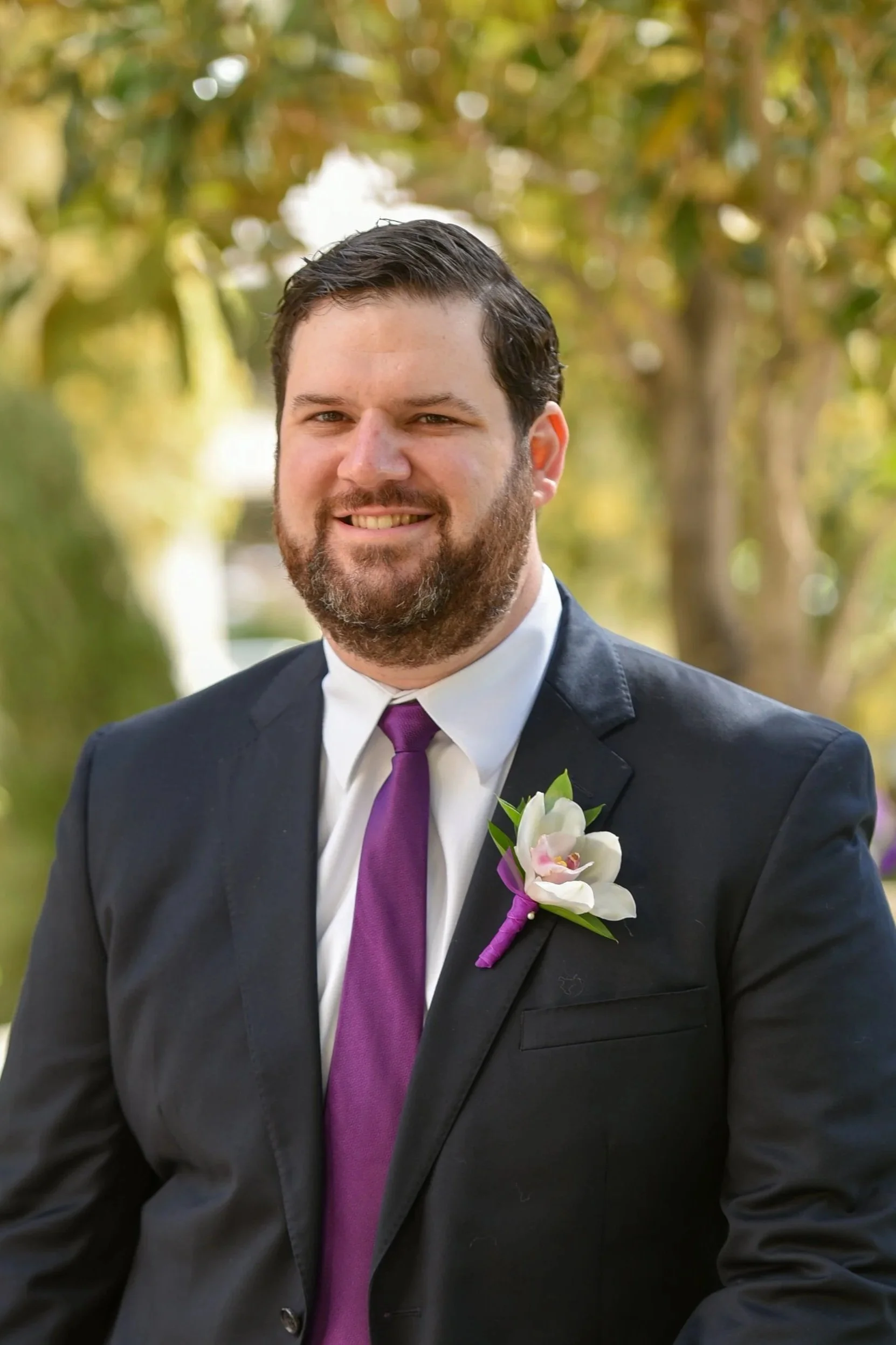 A man with dark hair, a beard, and a smile, wearing a black suit, white shirt, purple tie, and a boutonniere with white and pink flowers, standing outdoors with trees in the background.