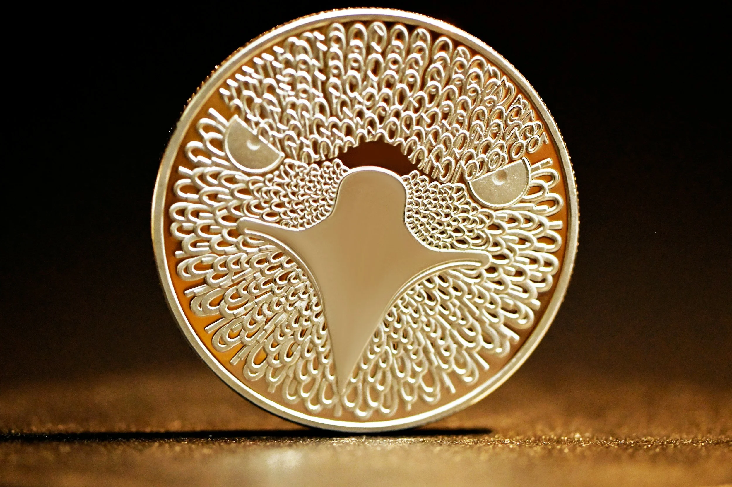 A gold-colored coin with an intricate design of a hummingbird surrounded by patterns and two small semicircular shapes.