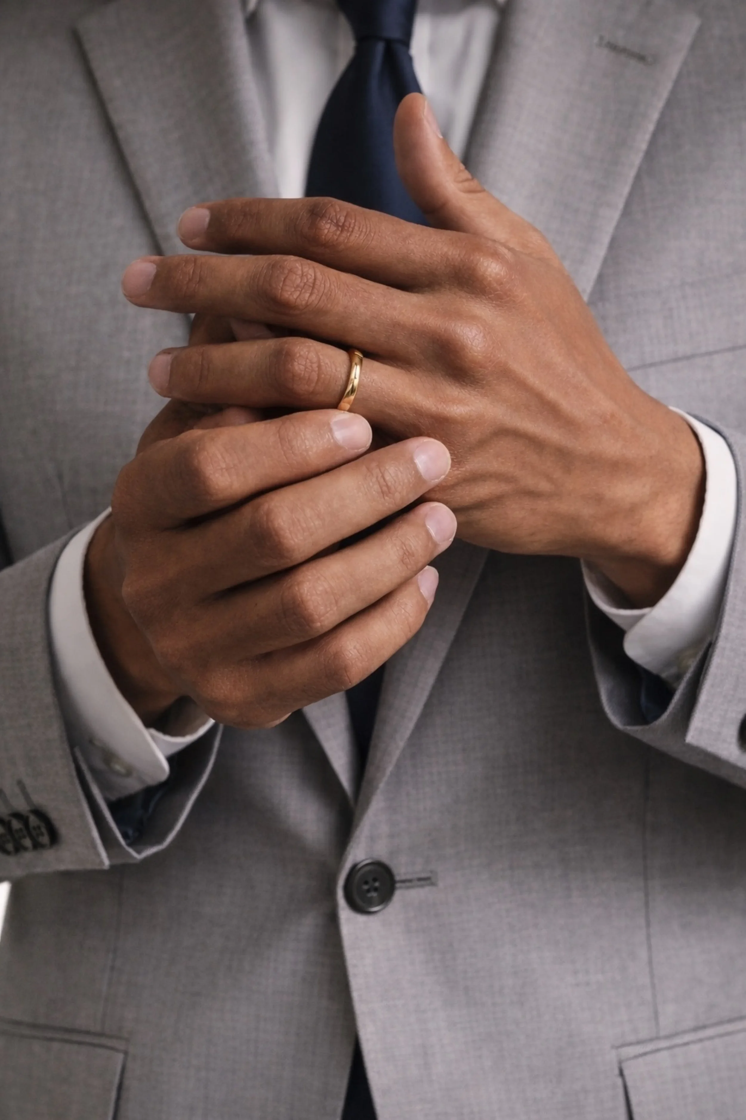 Close-up of a person in a light grey suit with a white shirt, wearing a wedding band, with their hands clasped.