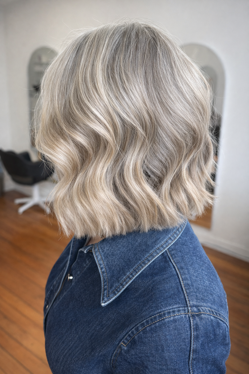 Back of a woman with shoulder-length wavy blonde hair wearing a denim jacket in a salon or similar setting.
