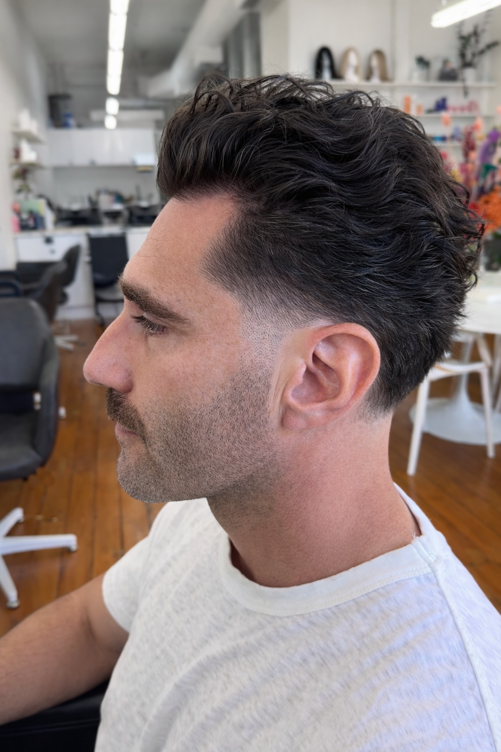 Side profile of a man with dark, wavy hair styled short on the sides and longer on top, sitting in a salon or barbershop.
