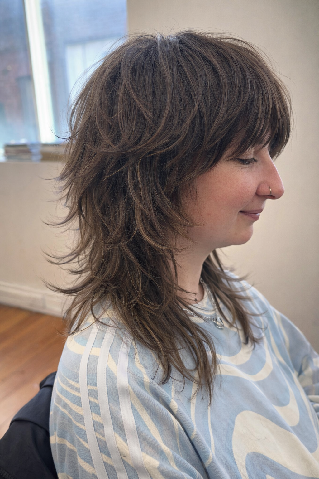 Profile of a woman with layered brown hair, wearing a nose ring and a light blue and white striped top, sitting indoors with a window in the background.
