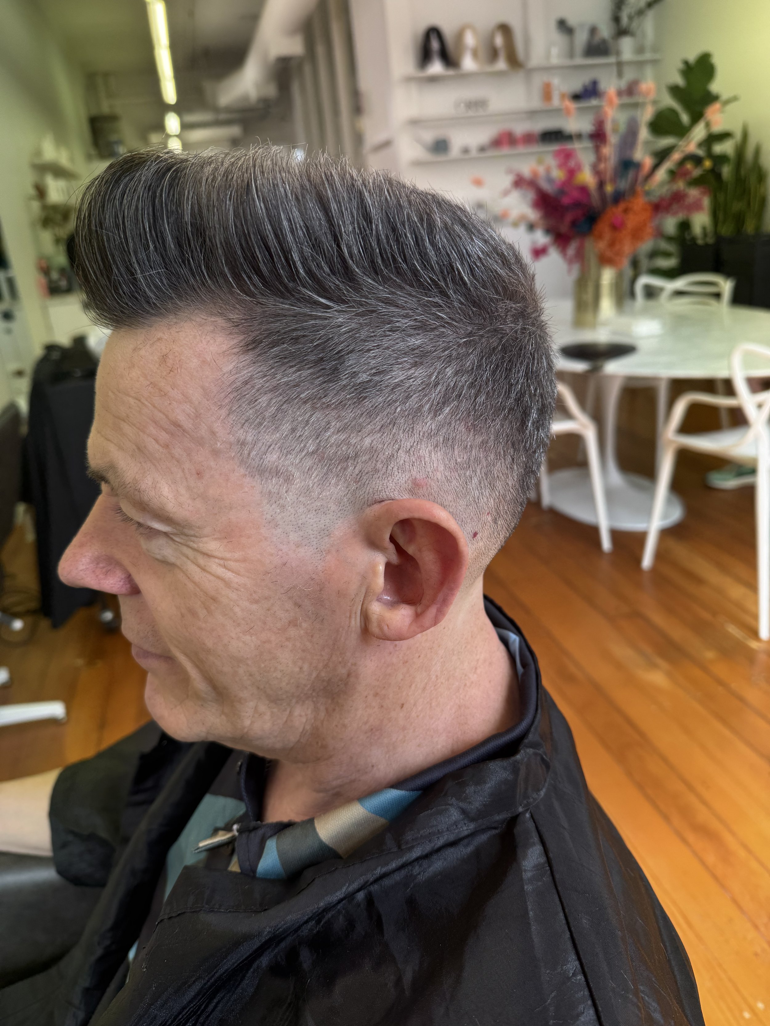 A man with a fresh high fade haircut, featuring dark hair on top and a gradual fade on the sides and back, sitting in a salon chair inside a brightly lit salon with wooden floors, white chairs, a table, and shelves with decorative items and a large f