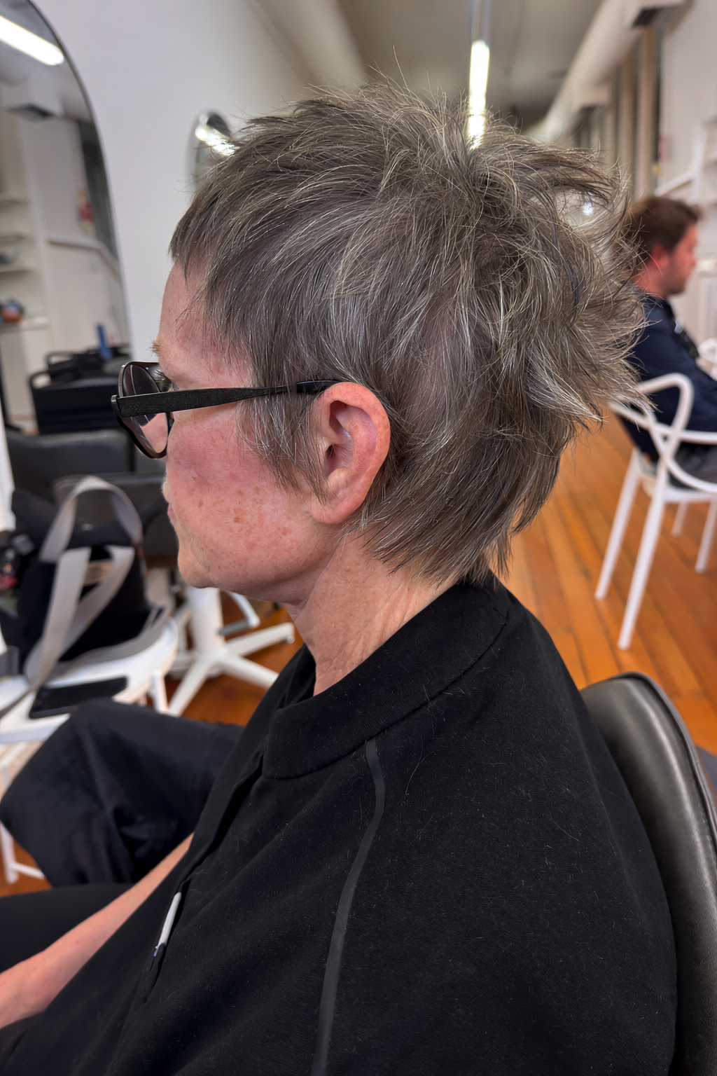 Side view of a woman with short, gray, and layered hair wearing black glasses, sitting in a salon or barbershop.