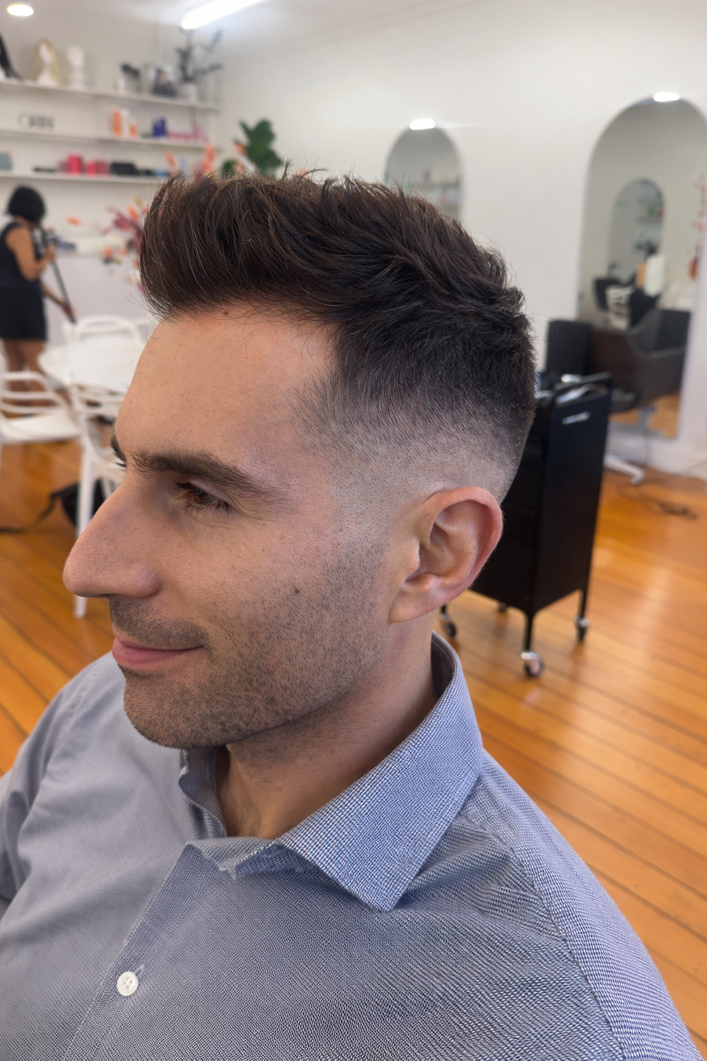 Young man with a fresh fades haircut seated in a salon, smiling with a gray button-down shirt, wooden floors, shelves with hair products, and a stylist in the background.