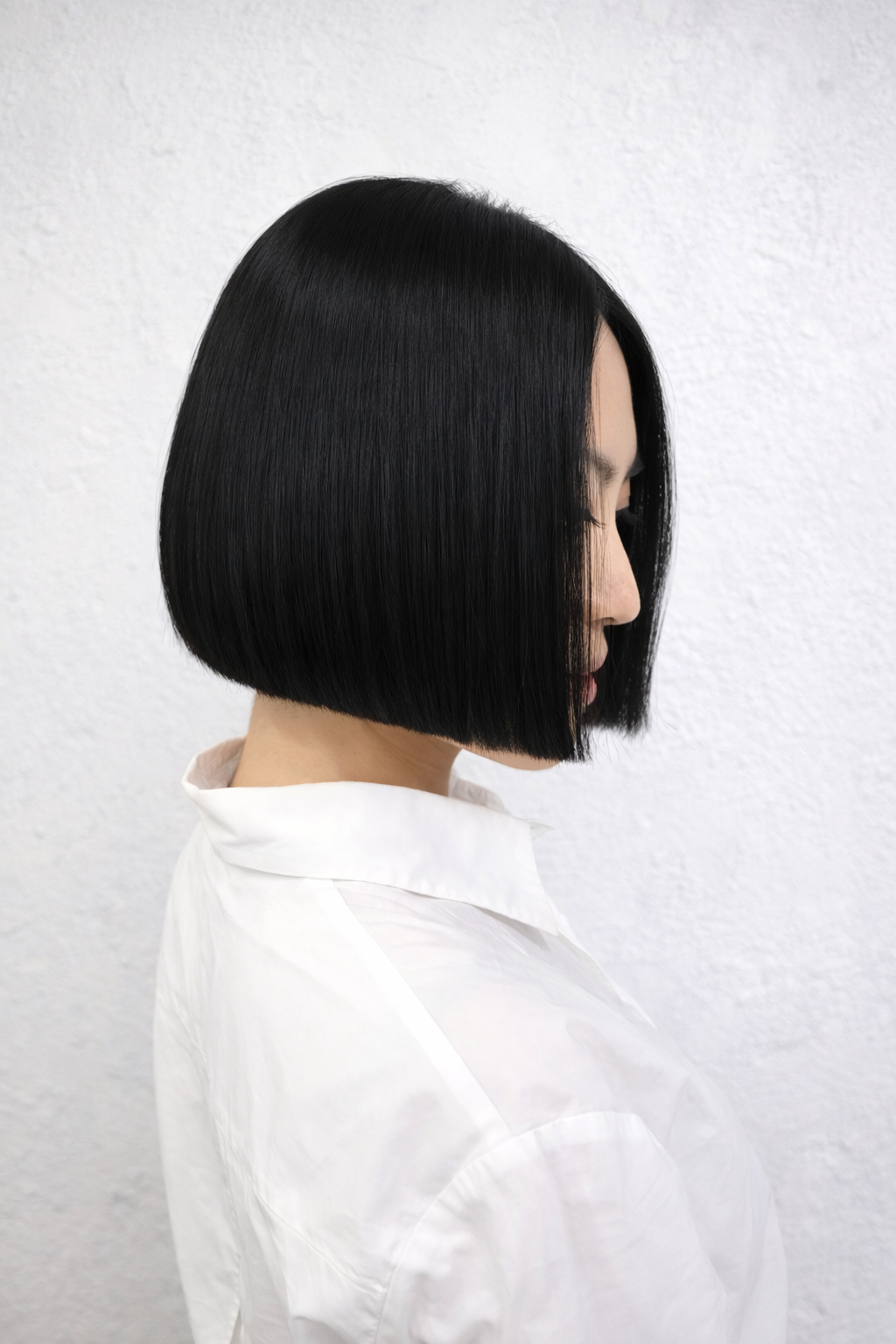 Side profile of a woman with a sleek, straight black bob haircut wearing a white blouse against a white textured wall.