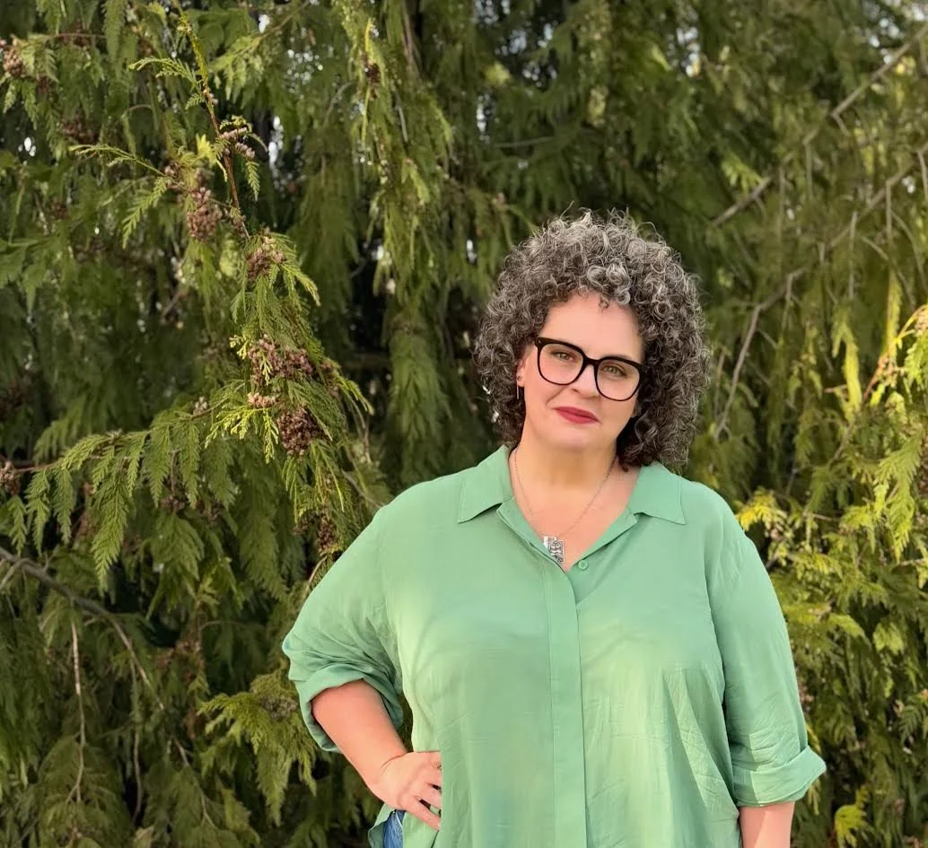 A woman with curly gray hair and glasses wearing a green shirt, standing outdoors in front of green trees.