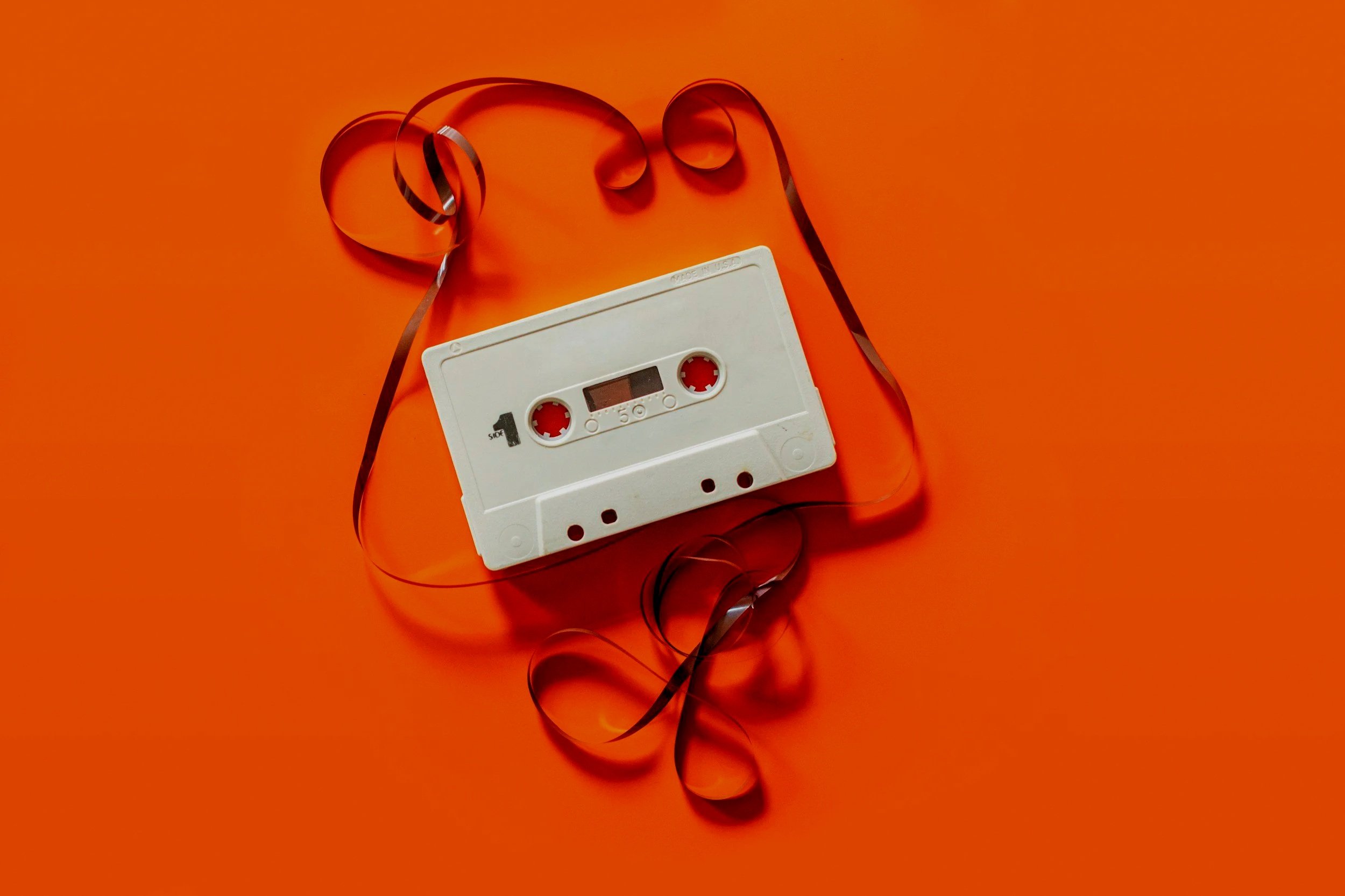 A white cassette tape with black spools and a small rectangular window, placed on an orange background, with black and orange curled ribbon around it.