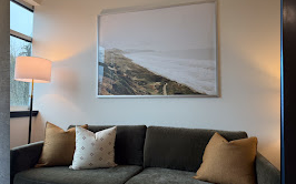 Living room with a dark sofa, beige and white pillows, a painting of a coastal landscape on the wall, a tall floor lamp, and a lamp with a beige shade.