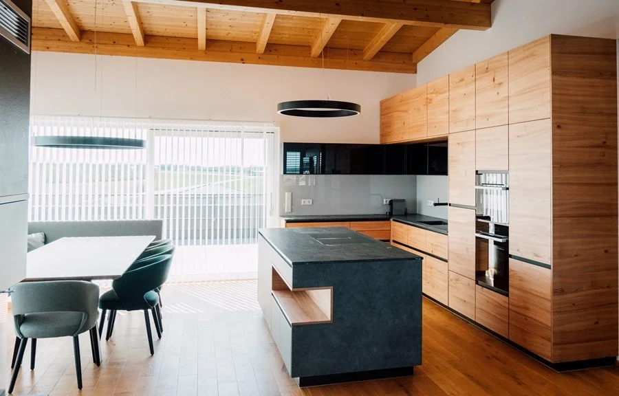 Moderne Küche mit Holzschränken, schwarzem Kochfeld, Esstisch mit Stühlen, große Fenster mit vertikalen Jalousien, Holzboden und Holzbalkendecke.