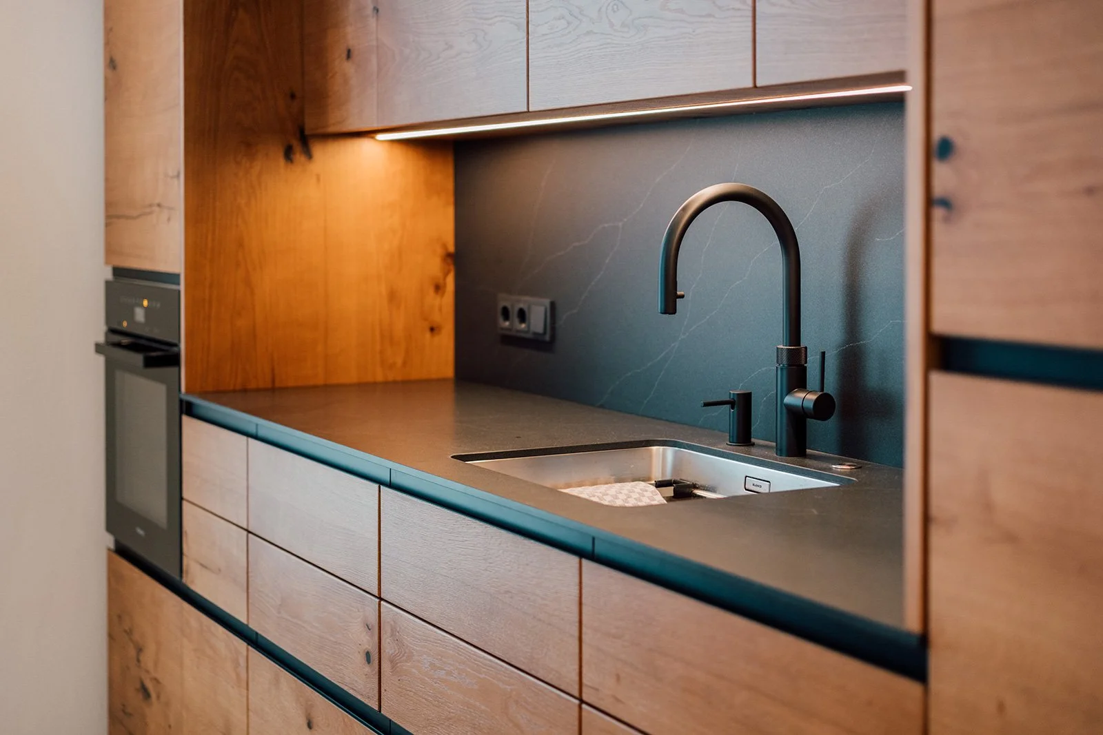 Moderne Küchenschrank mit schwarzer Spüle und schwarzer Armatur, Holzfronten, Backofen, graue Arbeitsplatte und Wand mit Steckdosen.