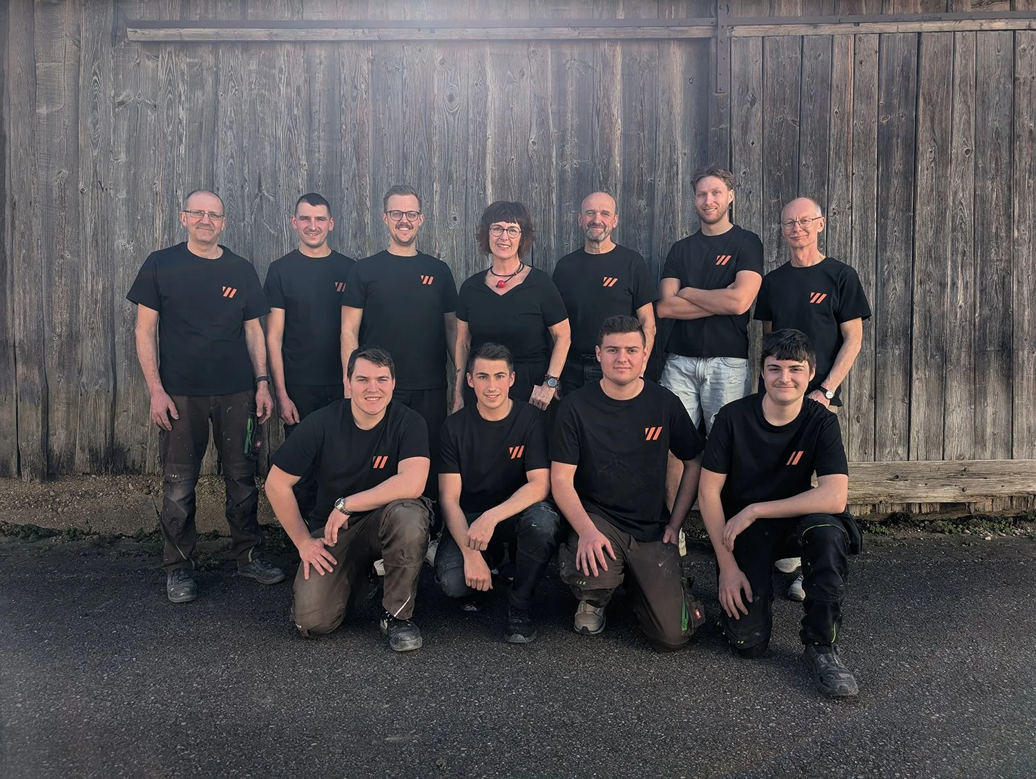 Gruppenfoto von zehn Menschen, die alle schwarze T-Shirts mit einem grauen und orangefarbenen Logo tragen, vor einer Holztafel auf einem Asphaltweg.