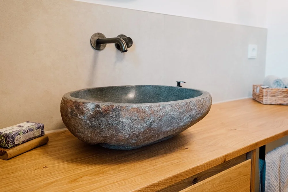 Ein runder, aus Stein gefertigter Waschbecken auf einem Holz-Waschtisch, mit Armatur an der Wand.