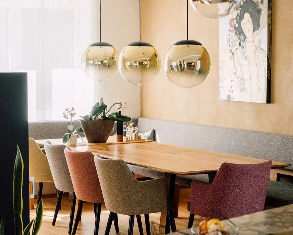 Ein Esszimmer mit einem langen Holztisch, bunten Stühlen, einer Pflanzenvase in der Mitte, hängende Lamellenleuchten, Wandkunst und einem Fenster mit Vorhängen.