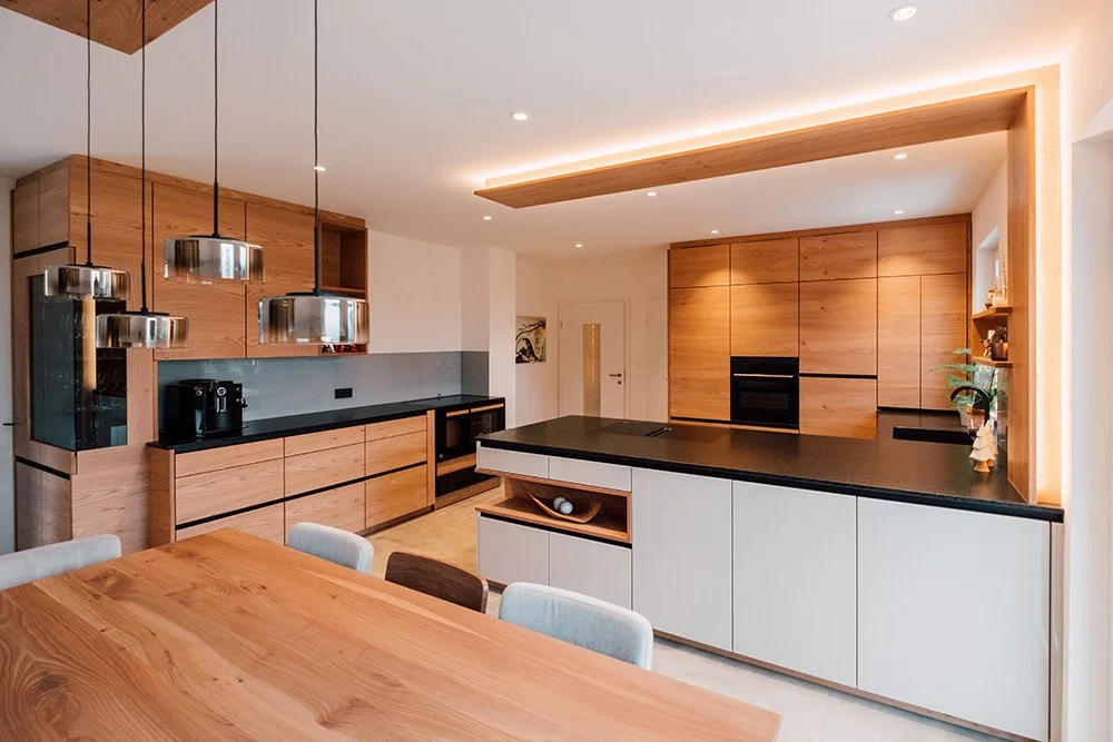 Moderne Küche mit Holzschränken, schwarzen Arbeitsplatten und einer weißen Insel, Hängelampen über dem Essbereich, kleine Regale mit Dekorationen, viel natürlichem Licht