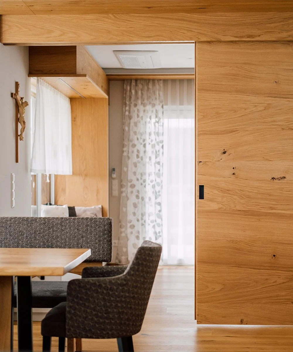 Innenansicht eines Wohnzimmers mit Holzverkleidung, einem Essbereich mit dunklen Stühlen, einem Sofa und Gardinen vor einem Fenster.
