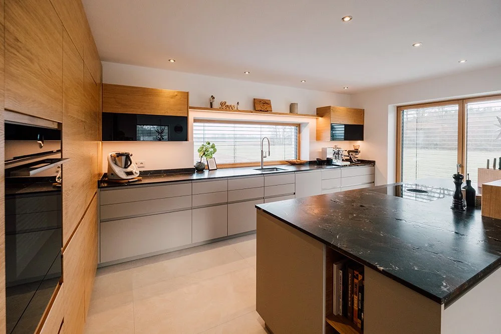 Moderne, helle Küche mit Holz- und grauen Schränken, einer schwarzen Steinplatte, großen Fenstern und Pflanzen.