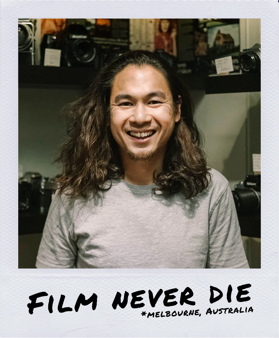 Portrait of a smiling man with long hair wearing a gray t-shirt, standing in front of a display of cameras and photography equipment.