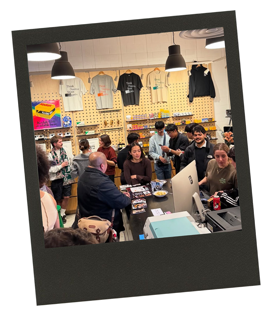 Group of people inside a retail store, some are shopping, others are chatting, and a few are working at the checkout counter. Shelves with various items and t-shirts are visible in the background.