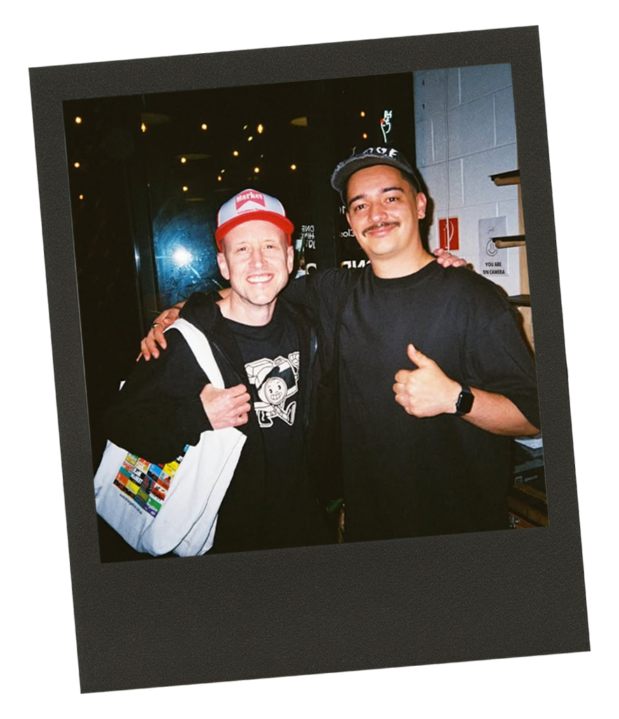 Two men smiling and posing together indoors at night. One man is wearing a black hoodie, a red and white cap, and carrying a colorful tote bag. The other man is wearing a black shirt and a gray cap. They have their arms around each other and are giving thumbs up.